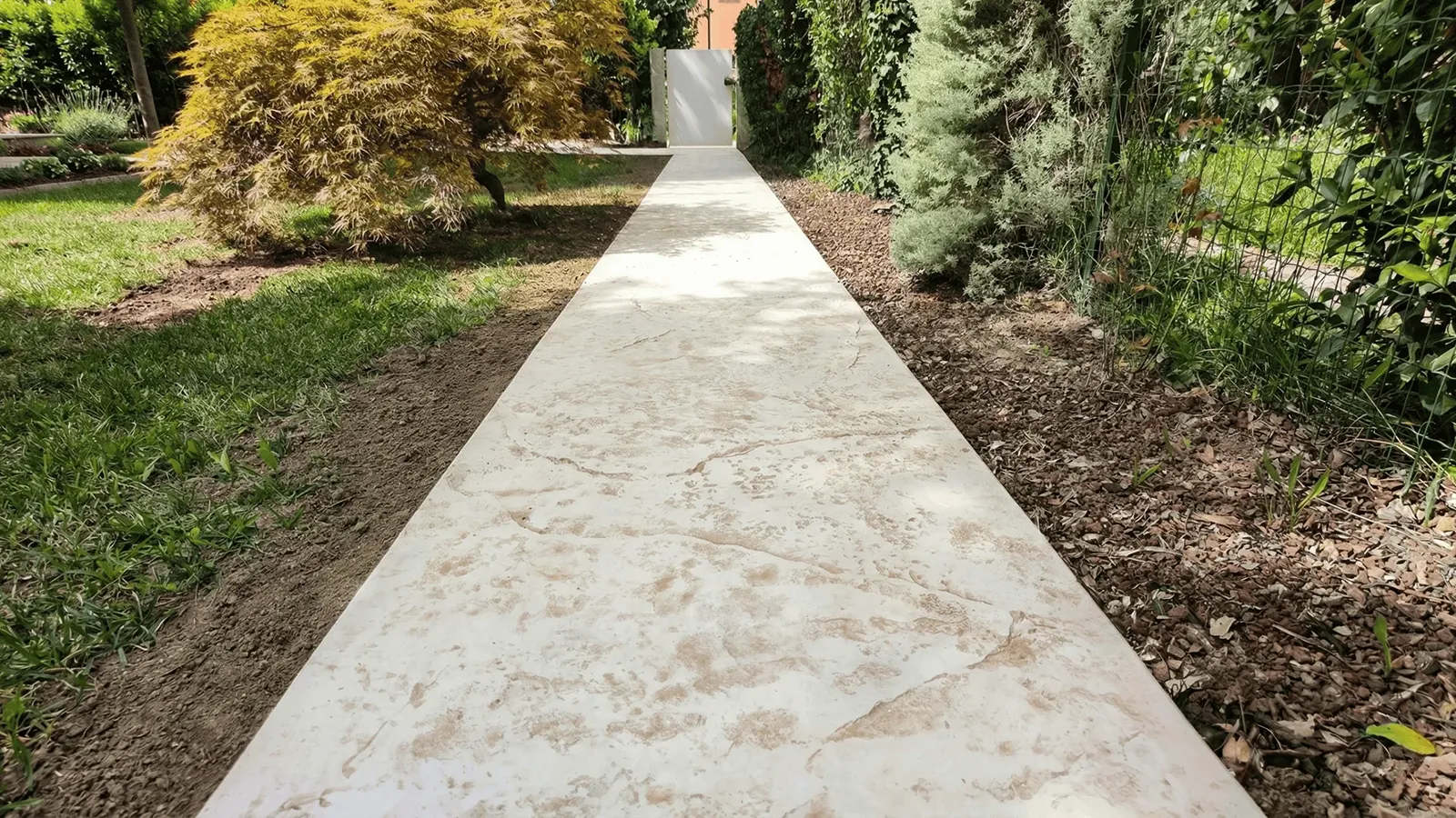 Vialetto in cemento stampato chiaro effetto pietra che attraversa un giardino con prato, aiuole e alberi ornamentali.