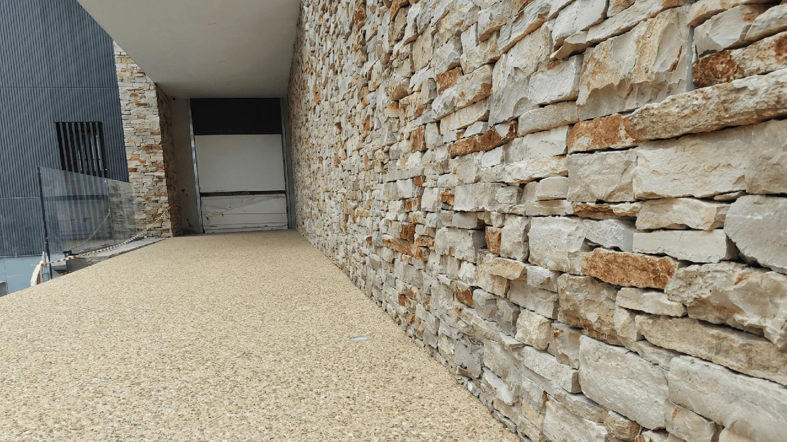 Passerella esterna con pavimento in ghiaino lavato chiaro affiancato da parete in pietra a spacco.