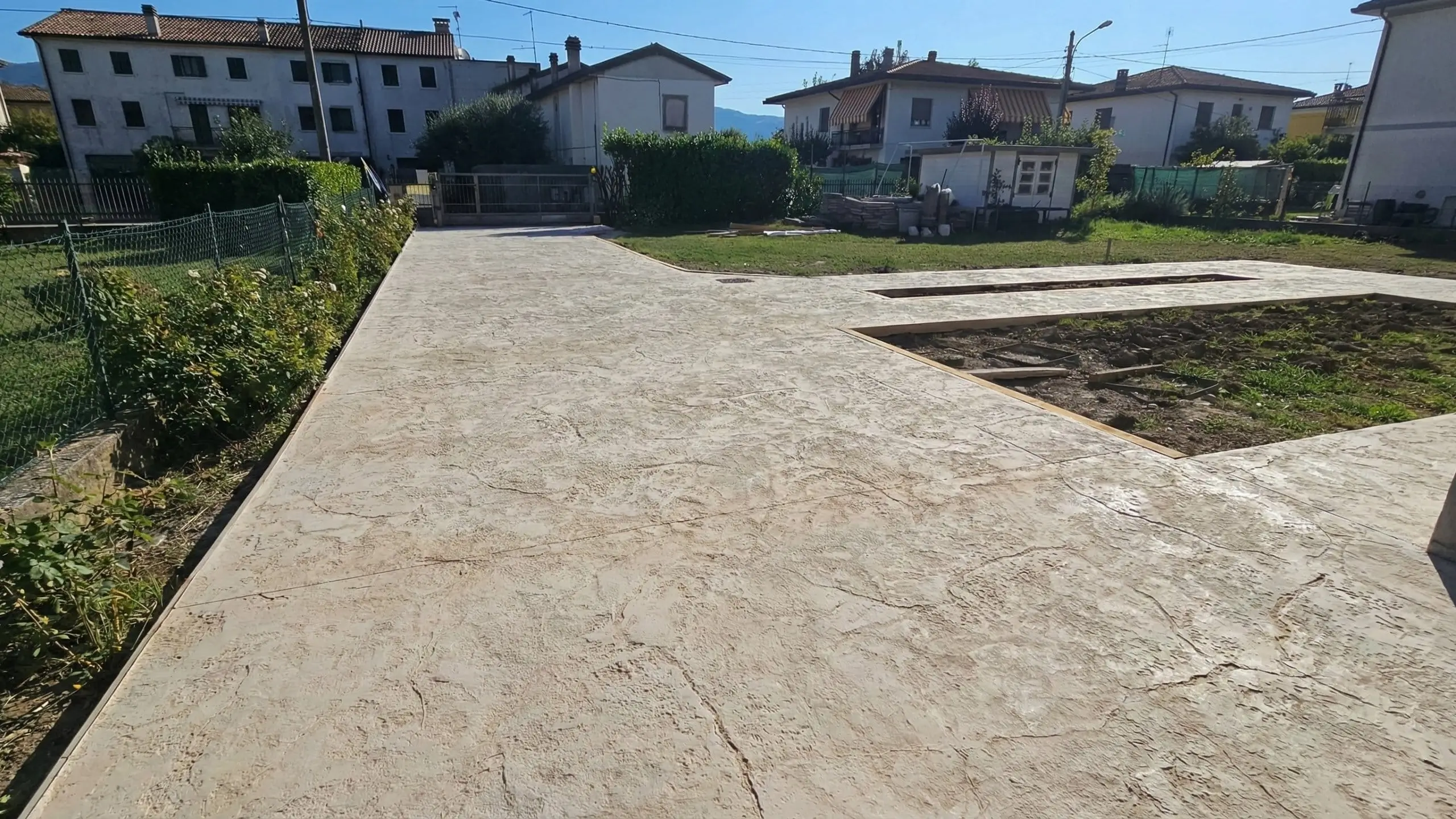 Piazzale in calcestruzzo stampato effetto sasso durante ristrutturazione: pavimentazione grigia drenante, giardini recintati e abitazioni vicine.